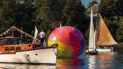 Bateau décoré de drapeaux, avec un homme debout à bord, naviguant près d'un grand ballon multicolore sur l'Erdre, pendant les Rendez-vous de l'Erdre, sous un ciel bleu. 