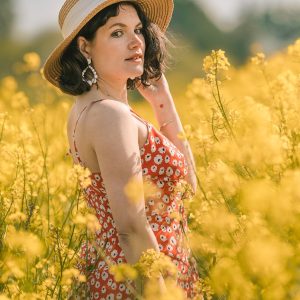 Portrait d'une jeune femme portant un chapeau de paille et une robe rouge à motifs floraux, se tenant au milieu d'un champ de colza en pleine floraison, regardant vers la caméra avec une expression sereine sous une lumière naturelle douce.