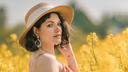 Portrait d'une jeune femme portant un chapeau de paille et une robe rouge à motifs floraux, se tenant au milieu d'un champ de colza en pleine floraison, regardant vers la caméra avec une expression sereine sous une lumière naturelle douce.