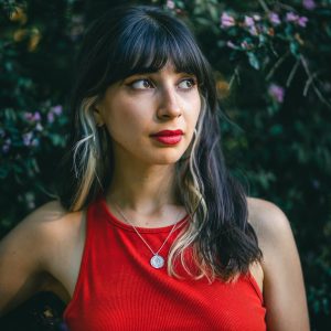 Portrait d'une jeune femme avec une expression pensive, portant un débardeur rouge et un collier avec un pendentif rond argenté, se tenant devant un fond de feuillage vert et fleuri.