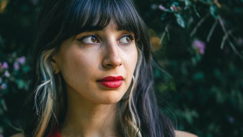 Portrait d'une jeune femme avec une expression pensive, portant un débardeur rouge et un collier avec un pendentif rond argenté, se tenant devant un fond de feuillage vert et fleuri.