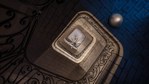 Vue en contre-plongée d'un escalier carré en pierre avec une rampe en fer forgé décorée, menant vers une ouverture lumineuse où l'on aperçoit un lustre moderne. 