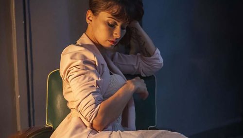Portrait d'une femme assise dans un fauteuil vert, portant un tailleur rose clair et des bottines noires à talons, avec une expression pensive. 