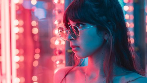 Portrait d'une jeune femme aux lunettes rondes près d'un jeu de fete foraine illuminé aux néons roses et bleus dans un style des années 80, avec une expression pensive.