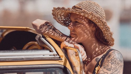 Portrait vintage de Danaë, portant une robe fleurie jaune et un chapeau de paille. Elle est appuyée sur une deux chevaux jaune et derrière elle on apperçoit la grue jaune de Nantes.