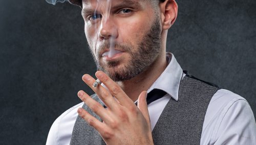 Homme fumant une cigarette, avec une chemise blance, une casquette et un gilet, pour un look insporé de la série Peacky Blinders. 