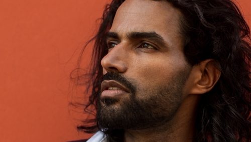 Portrait d'un homme barbu à la peau mate et aux cheveux long devant un mur orange.