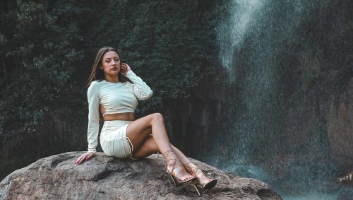 Jeune femme en 2 pièce blanc, assise sur un rocher devant une chute d'eau