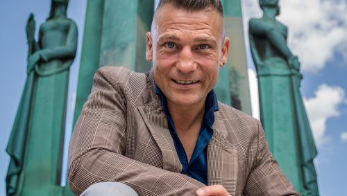 Portrait d'un homme souriant, portant un blazer à carreaux marron et une chemise bleue, agenouillé devant une structure architecturale verte flanquée de statues de femmes : le monument des 50 otages