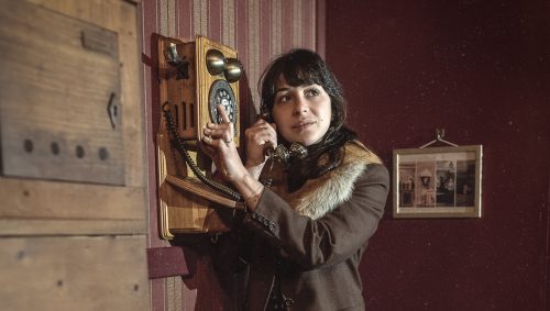 Portrait en éclairage studio d'une femme sur un ton vintage, parlant à un téléphone mural ancien, portant un tailleur marron avec une bordure de fourrure autour du cou et des escarpins vert foncé à talons. Elle a une expression pensive. Papier peint à rayures verticales beiges et bordeaux en arrière-plan. 