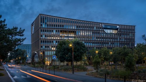 La Maison de l'Innovation à Nantes, photographiée à l'heure bleue, présente une façade moderne avec de hautes fenêtres et une section arrondie distinctive. Des traînées lumineuses rouges indiquent la circulation automobile sur une rue bordée d'arbres, éclairée par des lampadaires. Un bâtiment plus petit est visible à l'arrière-plan. 