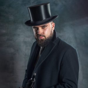 Un homme barbu vêtu d'un manteau sombre et d'un haut-de-forme tient un parapluie fermé et regarde l'appareil photo dans ce portrait dramatique réalisé en studio sur un fond sombre texturé.