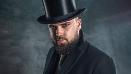 Un homme barbu vêtu d'un manteau sombre et d'un haut-de-forme tient un parapluie fermé et regarde l'appareil photo dans ce portrait dramatique réalisé en studio sur un fond sombre texturé. 
