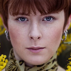 Jeune femme rousse entourée de fleurs de mimosa, regard direct.