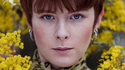 Jeune femme rousse entourée de fleurs de mimosa, regard direct.
