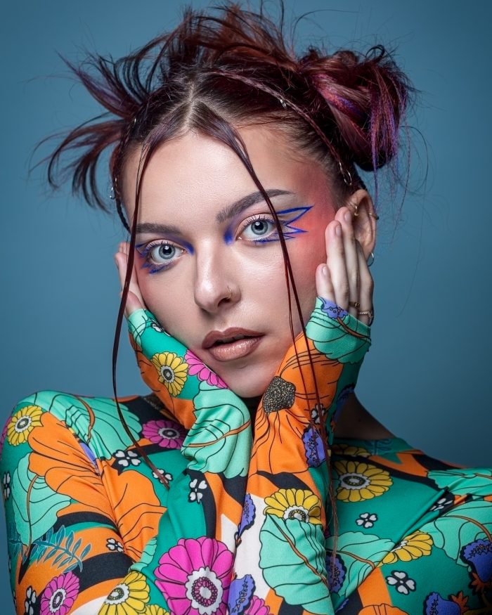Photographie portrait studio à Nantes : jeune femme avec un maquillage artistique bleu vif autour des yeux et les cheveux coiffés en chignons désordonnés, vêtue d'un top à motifs floraux colorés, sur un fond bleu uni, et ses mains posées sur ses joues, avec une expression douce.