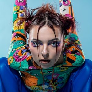 Jeune femme radieuse, vêtue d'un top à fleurs vive, les mains levées dans une pose espiègle. Son chignon désordonné et son regard captivant vous attirent, capturant l'essence d'une beauté vive, créée en studio à Nantes.