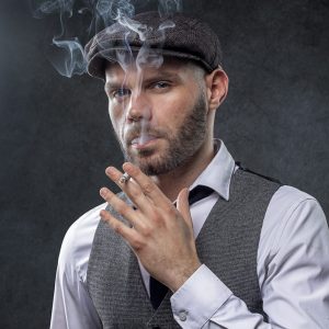 Cette image inspirée de Peaky Blinders représente un homme coiffé d'une casquette plate, vêtu d'une chemise et d'un gilet, fumant une cigarette, la fumée s'élevant au-dessus de sa tête sur fond sombre.