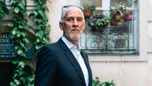 Portrait d'un homme mûr aux cheveux gris et à la barbe, portant un costume sombre sur une chemise claire, regardant directement la caméra avec une expression sérieuse, devant un bâtiment recouvert de verdure.