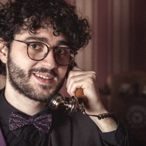 Jeune homme souriant avec des cheveux bouclés, portant des lunettes rondes et un noeud papillon à motifs, tenant un téléphone rétro à l'oreille dans un escape game à Nantes.
