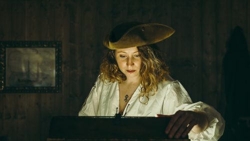 Dans un escape game une jeune femme en costume de pirate, concentrée et penchée sur un coffre illuminé par une lumière mystérieuse. 