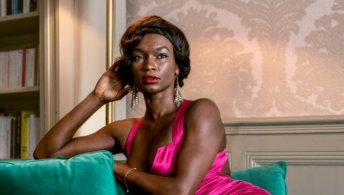 Portrait studio à domicile ou location airbnb ; photographie d'une femme assise sur un canapé vert émeraude, avec une expression confiante. Elle porte une robe fuchsia et des chaussures à talons hauts dorées. En arrière-plan, on aperçoit une bibliothèque et un papier peint à motifs délicats. Une lampe sur pied illumine la scène d'une lumière chaleureuse. 