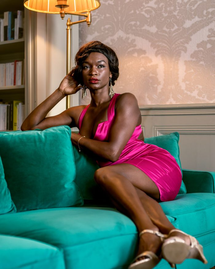 Portrait studio à domicile ou location airbnb ; photographie d'une femme assise sur un canapé vert émeraude, avec une expression confiante. Elle porte une robe fuchsia et des chaussures à talons hauts dorées. En arrière-plan, on aperçoit une bibliothèque et un papier peint à motifs délicats. Une lampe sur pied illumine la scène d'une lumière chaleureuse.