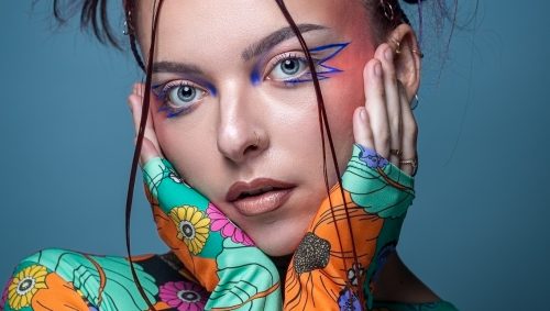 Portrait d'une femme avec un maquillage artistique bleu et rose inspiré d'Euphoria, portant un haut à motifs floraux colorés, adoptant une expression neutre. 