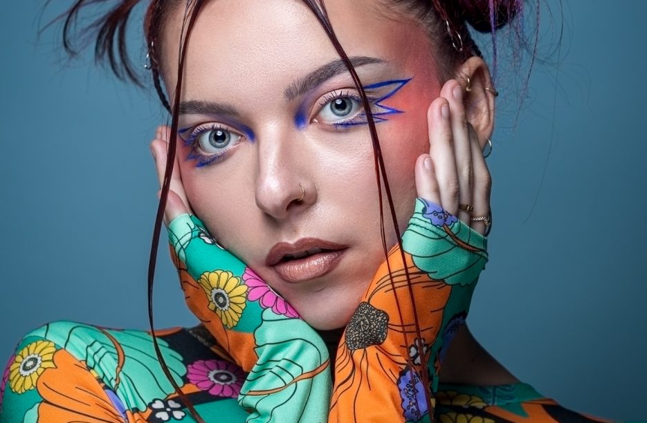 Portrait d'une femme avec un maquillage artistique bleu et rose inspiré d'Euphoria, portant un haut à motifs floraux colorés, adoptant une expression neutre.