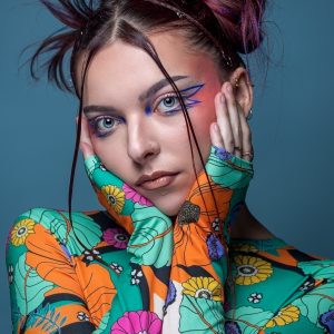  Jeune femme avec un maquillage inspiré de la série "Euphoria". Ses cheveux auburn décoiffés ont des reflets violets. Elle porte un haut à motifs floraux colorés et pose ses mains sur son visage. Elle porte un anneau dans le nez.