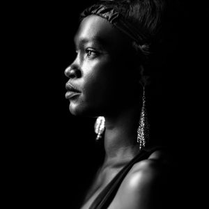 Portrait studio en noir et blanc d'une femme noire de profil, avec une expression sérieuse et un bandeau dans les cheveux, portant des boucles d'oreilles pendantes,