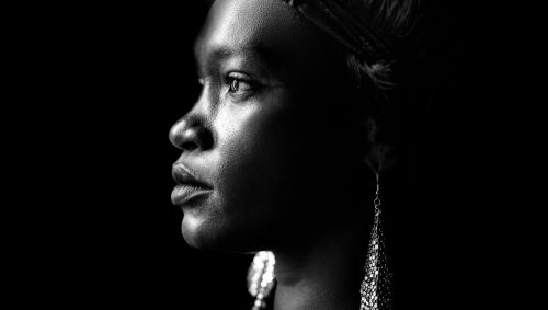 Portrait studio en noir et blanc d'une femme noire de profil, avec une expression sérieuse et un bandeau dans les cheveux, portant des boucles d'oreilles pendantes, 