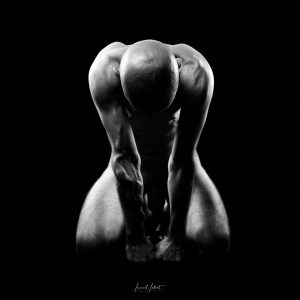 Portrait en noir et blanc d'un homme nu agenouillé, la tête baissée, dans un studio sur fond noir.