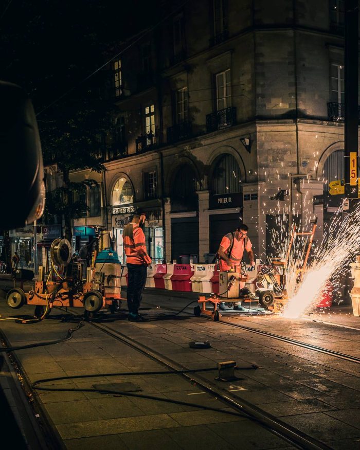 Travaux nocturnes sur une ligne de tramway, deux ouvriers en gilet orange entretiennent des rails, produisant des gerbes d'étincelles spectaculaires.