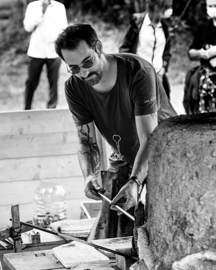 Un verrier concentré manipule une tige près d'un four, son visage reflétant l'attention portée à son métier d'art.