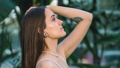 Portrait dans une serre d'une jeune femme brune, main dans la poche de son pantalon bleu turquoise et l'autre dans ses cheveux, regardant vers le haut d'un air pensif.