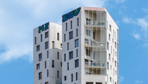Le Duo des Cimes à Nantes, un ensemble architectural moderne en béton blanc, se dresse sous un ciel bleu clair avec quelques nuages ; des graffitis colorés décorent le sommet des bâtiments.