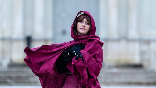 Portrait d'une jeune femme emmitouflée dans une cape bordeaux à capuche, devant un bâtiment blanc classique, un jour de neige. Elle croise ses mains gantées de noir sur sa poitrine et regarde l'objectif avec une expression neutre.