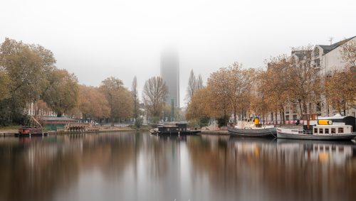 Brouillard sur l'Erdre à Nantes, décor automnal avec des arbres aux feuilles orangées et des péniches amarrées le long des quais. La tour Bretagne se dresse au loin, partiellement masquée par la brume. L'eau est lisse et reflète le paysage. 