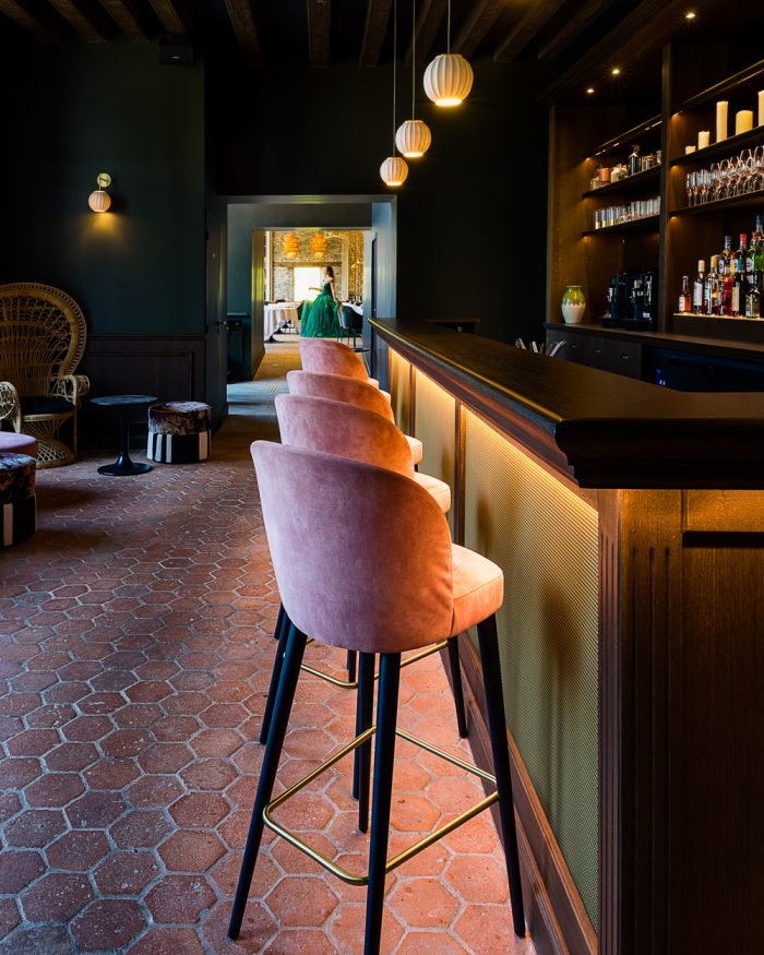Intérieur chaleureux d'un restaurant avec des tabourets roses devant un bar bien éclairé, un sol en terre cuite hexagonale et une femme en robe verte à l'arrière-plan.