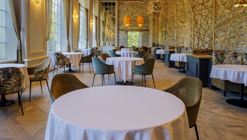 Intérieur de restaurant avec tables rondes nappées de blanc, chaises vertes et motif décoratif en osier au plafond. Le sol est en parquet clair. 