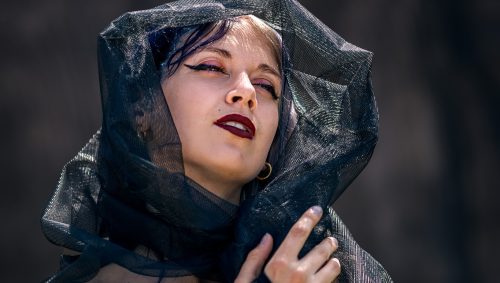 Portrait d'une jeune femme regardant vers le haut, un voile texturé noir sur la tête et les épaules, gant de résille, maquillage expressif. Elle a un tatouage sur son épaule dénudée.