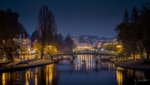 Vue nocturne de Nantes à l'heure bleue, avec le pont Saint-Mihiel enjambant l'Erdre, illuminé par des réverbères jaunes chaleureux qui se reflètent dans l'eau calme. 