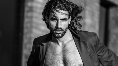 Portrait en noir et blanc d'un homme musclé, torse nu avec une veste de costume, regard sérieux et cheveux bouclés au vent.