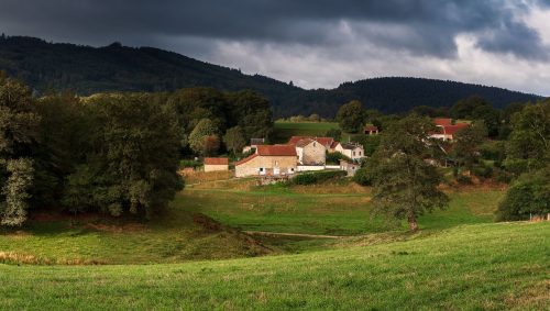 Paysage verdoyant de la campagne française avec quelques maisons et une forêt en arrière-plan sous un ciel nuageux menaçant. 