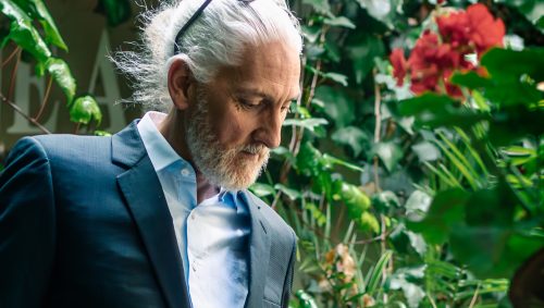 Portrait d'un homme d'âge mûr avec une queue de cheval grise, portant un costume bleu foncé et examinant de près une fleur rose.
