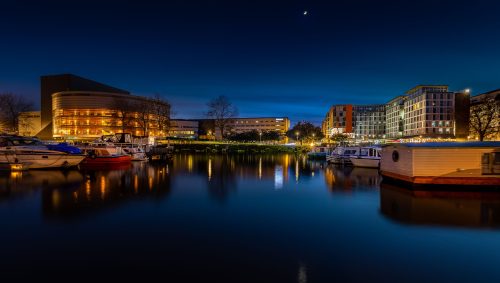 Une vue nocturne du canal Saint-Félix à Nantes, avec le centre des congrès illuminé et des bateaux amarrés le long des quais, sous un ciel bleu profond avec un fin croissant de lune. 