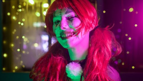 Portrait d'une personne déguisée pour Halloween en poupée effrayante, avec perruque rousse, maquillage pâle avec des cicatrices et un sourire inquiétant. Des lumières colorées vertes, jaunes et mauves illuminent l'arrière-plan.
