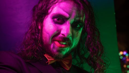 Un homme sourit, maquillé comme Jigsaw pour Halloween, avec des spirales rouges sur les joues et portant un nœud papillon orange, le tout éclairé en violet et vert.