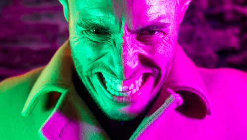 Portrait d'un homme arbora,nt le sourire du Joker pour Halloween, éclairé en vert d'un côté et en rose vif de l'autre, affichant un rictus menaçant.